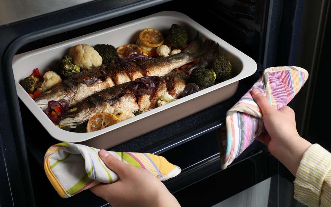 Woman taking out baking tray with sea bass fish and vegetables from oven, closeup